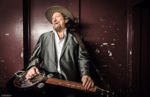 Jerry Douglas wearing a grey suit and a Stetson hat with his trusty Dobro guitar in his lap.