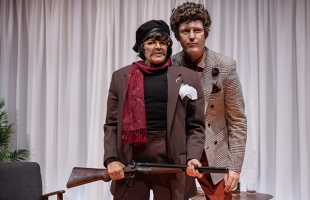MASTERCLASS poster, Adrienne Truscott, dressed as a man, with a moustache, and over-sized jacket, is holding a rifle and standing in front of Feidlim Cannon, who has a curly wig and a chequered blazer.