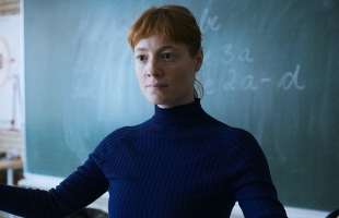 A woman with red hair, the protagonist of The Teacher's Lounge, standing in front of a blackboard. She is wearing a dark blue polo neck jumper, and has red hair.