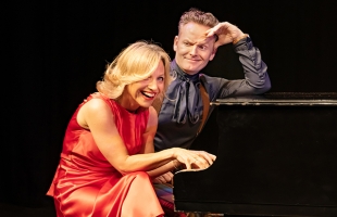 Liza Pulman in a red dress and Joe Stilgoe in a grey shirt and suspenders. They are both sat at a piano, Liza with her fingers on the keys, laughing, and Joe with a quizzical look on his face and his hand cradling his forehead in an