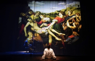 A man in a white shirt sitting on stage, his back to us, staring up at a huge depiction of Raphael's