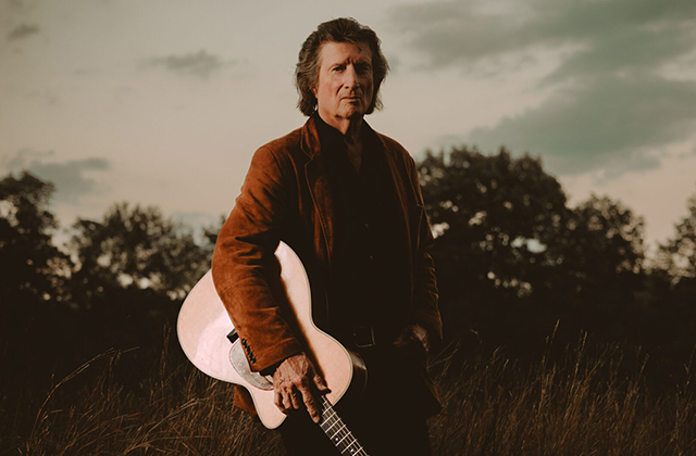 Chris Smither holding an acoustic guitar in one hand. He is wearing a thick brown-orange jacket and his hair is swept back. Behind him there are trees and a dusky sky.