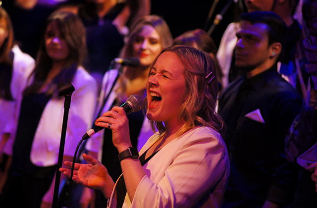 A photograph of a member of Dublin Gospel Choir, she is wearing a white suit and singing energetically into the microphone. The rest of the choir stands behind her.