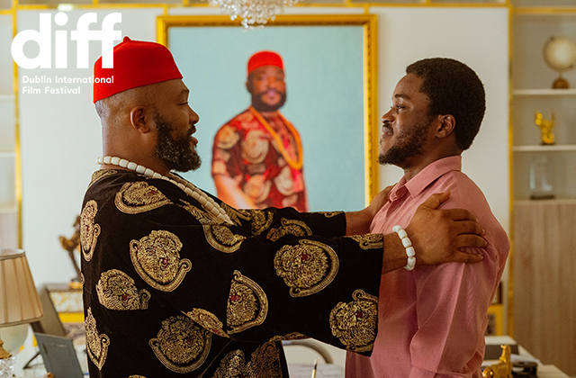 Photograph of a bearded middle aged man in a red fez-like hat and resplendent gold and black robes, holding a younger man in a pink shirt by the shoulders. Both appear to be happy. In the background there is a portrait style painting of a man dressed similarly to the man on the left, possibly himself or a relative of his.