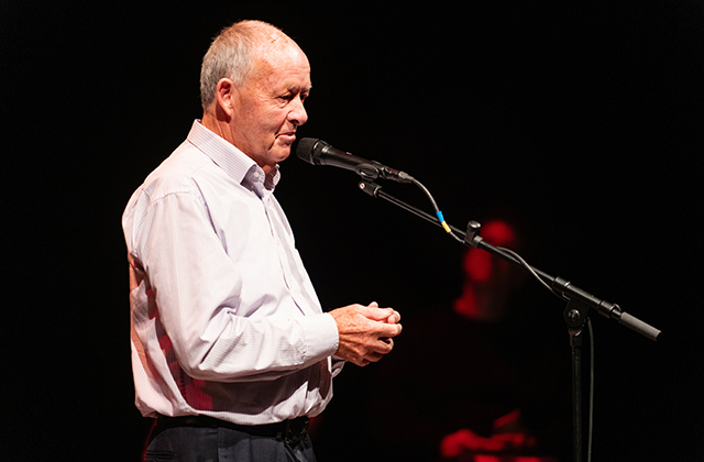 An older man in a white shirt, telling a story on stage.