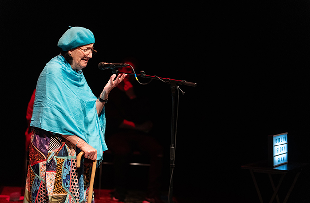 An older woman wearing a blue beret and shawl, telling a story on stage.