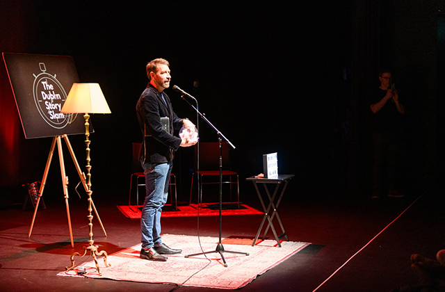 Host Colm O'Regan in a black jacket and jeans, presenting Dublin Story Slam on stage.