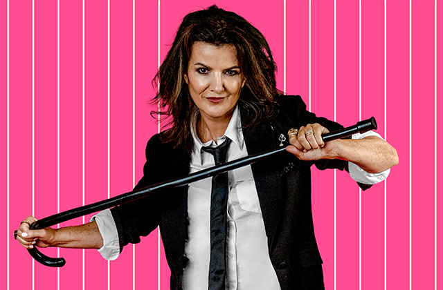 Deirdre O'Kane in a back suit with white shirt and black tie, holding a black cane in an