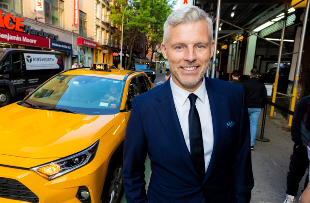 Des Bishop stands in a suit on a New York street, with a Yellow Taxi cab behind him