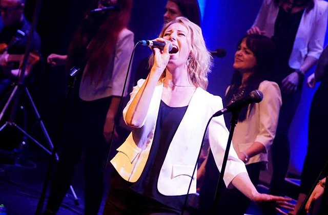 A photograph of a member of Dublin Gospel Choir, she is wearing a white suit and singing energetically into the microphone. The rest of the choir stands behind her.