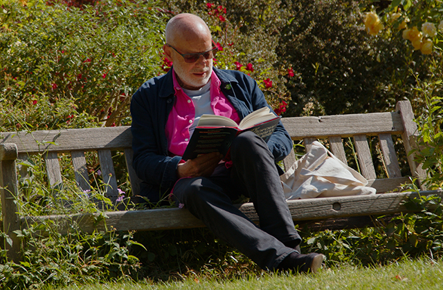 Brian Eno on a garden bench, reading