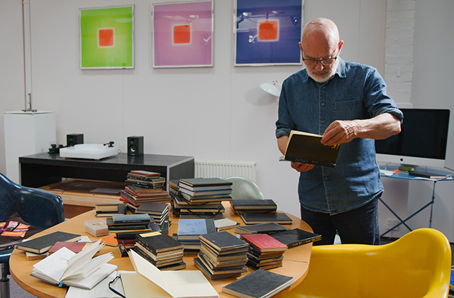 Brian Eno in an office, reading a notebook behind a cluttered desk