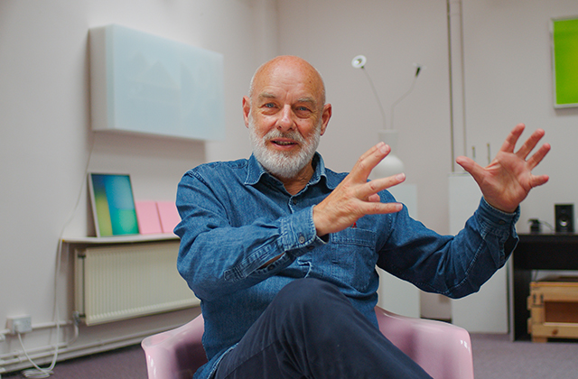 Brian Eno, sitting, explaining something using his hands