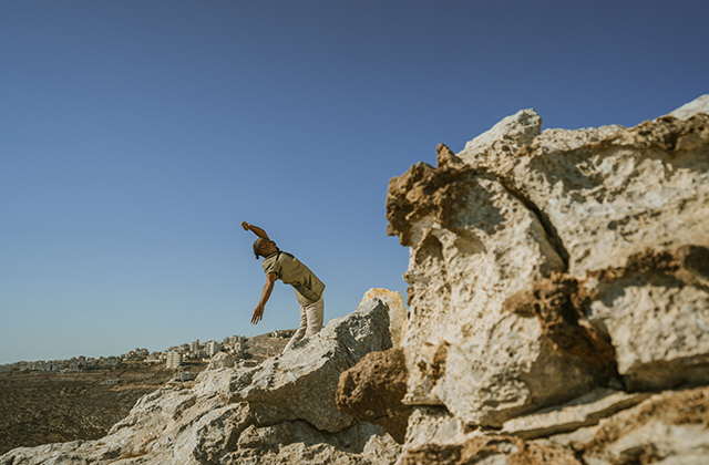 A dancer bending backwards, framed by craggy sand-coloured rocks and a bright blue sky.