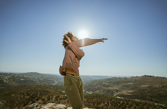 A dancer making a cross with his hands, backlit by the sun, with a view of the Palestinian countryside in the background.