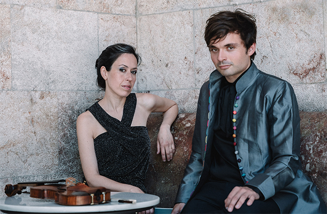 Alba Venura and Francisco Fullana seated next to one another against a stone wall. There is a violin on a nearby table. The two are dressed formally.