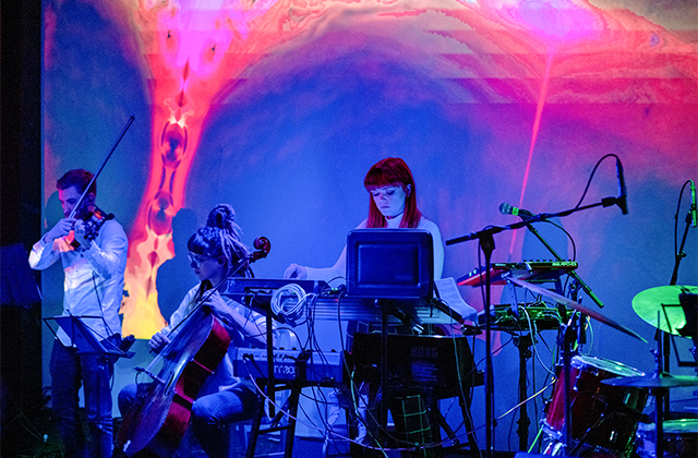 Blue-tinged photo of Glasshouse on stage performing, with an abstract backdrop. There is a man playing violin, a woman playing cello, and a woman at a synthesiser console.