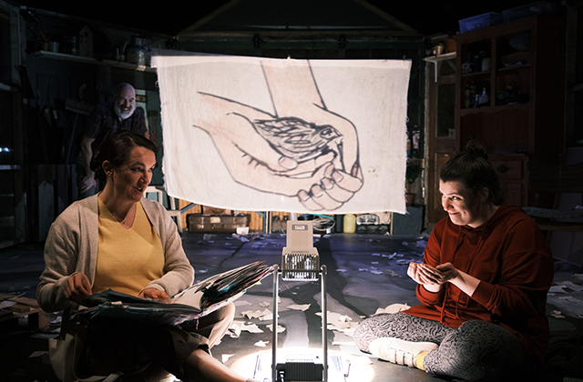 Grace Kiely and Eleanor Walsh, as Mam and Grace, sat on opposite sides of a overhead projector. A line drawing of a sparrow in cupped hands is projected on to a white sheet behind them.