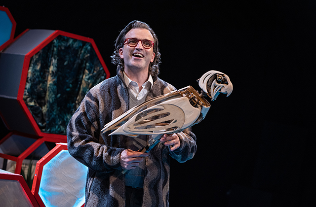 Paul Curley holding a wooden mechanical parrot, wearing a stripy robe, grey jumper and white collared shirt, with a big smile on his face and a glint in his eye.