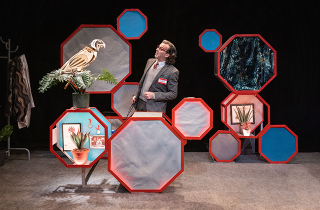 Paul Curley in a tartan jacket, beige jumper and red tie, speaking to a wooden mechanical parrot with a series of red-bordered octagons around them. Some of the octagons are behaving like furniture, holding objects such as houseplants. Others have blank grey faces.