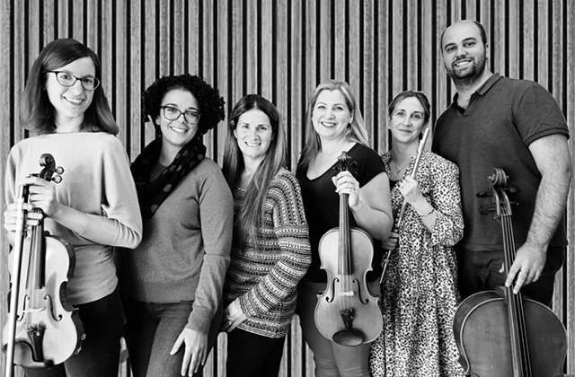 A black and white shot of six musicians against a background of wood panelling. All but one are women, and three hold string instruments.