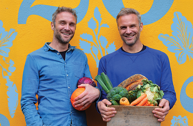 A photo of Dave and Steve Flynn, smiling, both wearing blue and holding various fruits and vegetables. Behind them is a yellow wall with brightly painted blue murals.