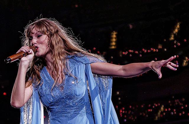 Taylor swift in a blue dress, singing into a microphone with her left arm outstretched. A stadium crowd with red lights aloft is out of focus behind her.