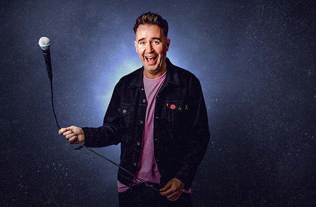 Jarlath Regan in a pink t-shirt and dark denim jacket, smiling widely and swinging a microphone cable.