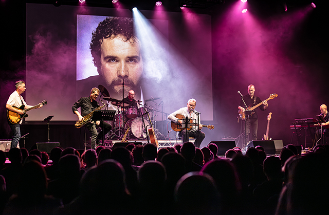 Stage shot of Ken Haddock and his band performing, with a large photo of John Martyn projected behind them.