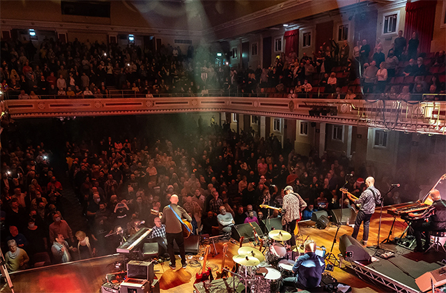 Overhead shot of Lindisfarne from behind, where we can see they are playing to a packed, multi-level auditorium.