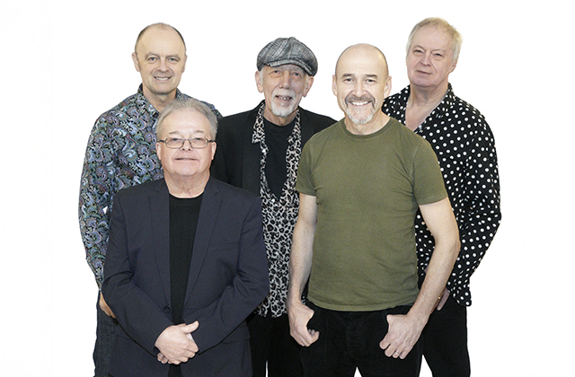 Five members of Lindisfarne stand together against a white background, they are looking towards the camera smiling, Rod Clements stands in the centre wearing a cap, shits and blazer.