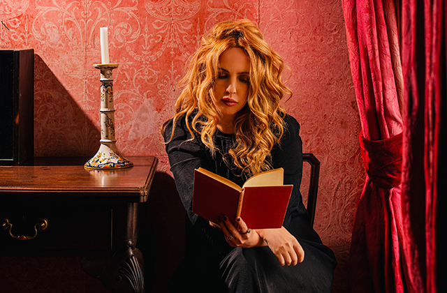 Lisa Lambe sits on a chair with her hair hanging in front of her face, reading a book with a red cover, in a room with red wallpaper, and a red velvet curtain hanging to her left. There is a table with an ornate candlestick to her right.