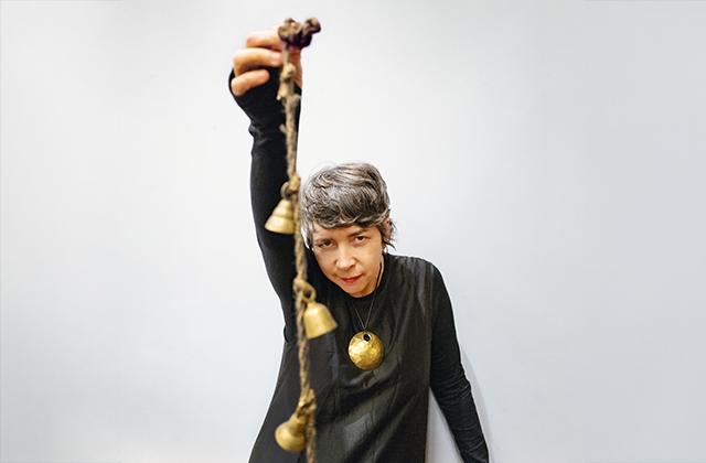 Lisa O'Neill in a long-sleeve grey top, with her right arm stretched above her head, holding a rope with small golden bells hanging off it.