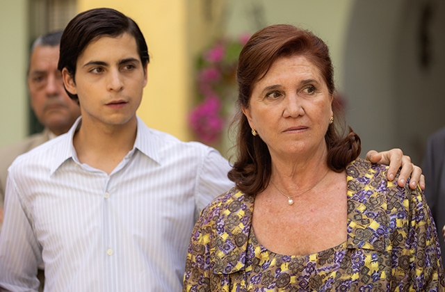 Still from Love & Revolution. A young man in a white shirt stands next to an older woman in a yellow and purple blouse. Both look suspicious.