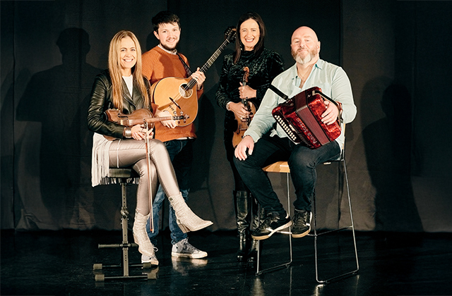 Yvonne Kane, MacDara Ó Faoláin, Liz Kane, and Derek Hickey pose on a black stage, with their instruments - fiddle, bouzouki, fiddle and accordion respectively.