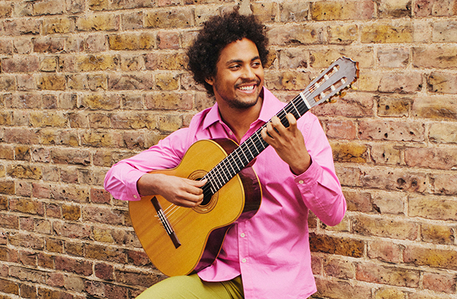 Plínio Fernandes, wearing a pink shirt and holding a guitar, leans against a brick wall, smiling