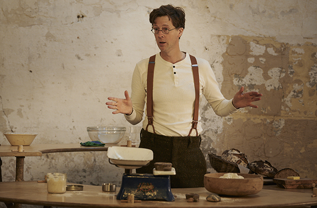 Manchán Magan at a weighing scales gesticulating as if explaining something. He wears suspenders and a white, longsleeve t-shirt.