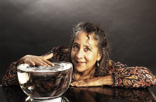 Marilyn Mazur leaning forward on a table, with a large bowl of water. Her hand is creating ripples in the water.