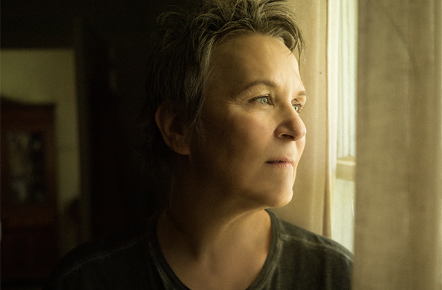 Mary Gauthier is pictured up close, she is standing in a room that appears to be a house, it is dark in the room and there is light appearing onto her face from the window which she is looking out of.