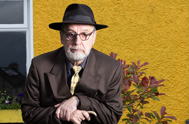 Michael Harding in a dark suit, yellow tie, trilby hat and round glasses. There is a yellow wall behind him.