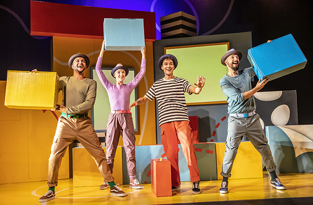 Human cast of the show appearing to be mid-song, dancing and holding large boxes on a colourful set.