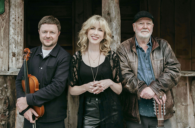 The Muireann Nic Amhlaoibh Trio standing in front of an old wooden building - left to right: Dónal O'Connor wearing a black shirt, fiddle in hand, Muireann Nic Amhlaoibh with her fingers interlaced, and Gerry O'Beirne in a leather jacket, his hands resting on the headstock of his guitar.