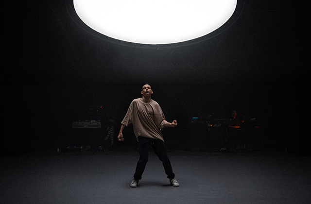 A solo male dancer in a baggy cream t-shirt dances under a huge white light.