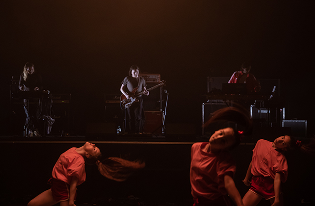 Young dancers whip their hair in foreground, behind three musicians play under dramatic spotlight.