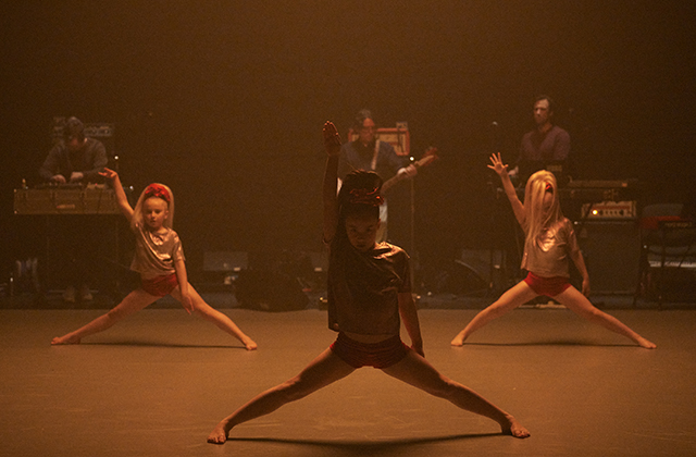 Three young girls stand in semi-splits, with one hand raised. Behind them, a band plays.