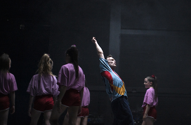 A male dancer is central, reaching up, wearing a multicoloured top and illuminated, he is surrounded by females in pink tops, and red shorts