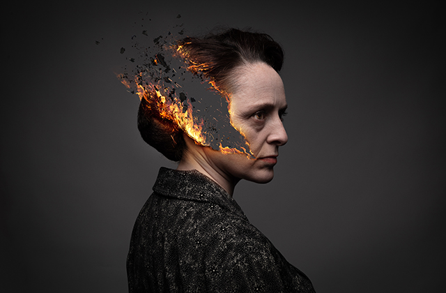 Mary Murray in profile, her face drawn and sombre, wearing all black, with a stylised streak of flame and ash burning through her hair, ear and cheek.
