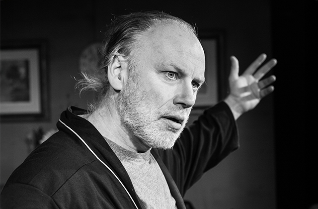 Dramatic black-and-white close-up of Don Wycherley in a dark robe, his left hand raised and an intense expression on his face.