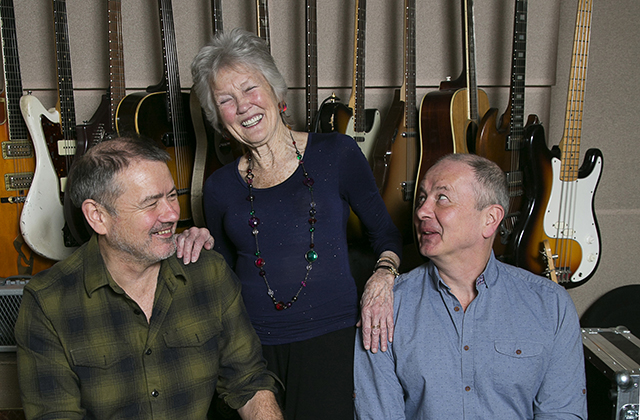 Peggy Seeger smiling widely, with her hands on the shoulders of her two sons as they look up at her. There's a wall of guitars hanging behind them.