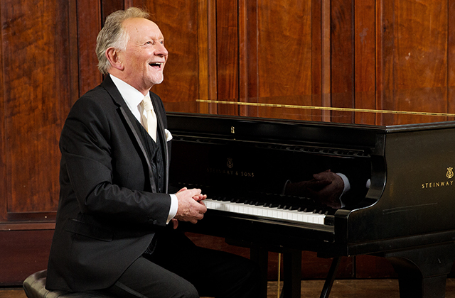 Phil Coulter sits at his piano smiling looking out into the distance. He is wearing a suit and appears to be in a rehearsal room.